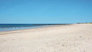 Una amplia playa arenosa con agua tranquila, un rompeolas rocoso y un faro a lo lejos.