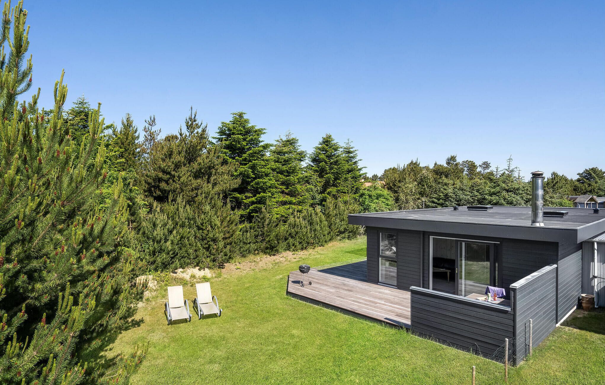 Ein modernes Haus mit einer Terrasse, einem Grill und einem großen Baum im Hintergrund.