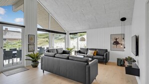 Un salon spacieux avec un grand canapé gris, un canapé plus petit agrémenté d’un coussin à motifs et une table basse. On y trouve deux plantes en pot, un tableau encadré accroché au mur et un luminaire suspendu.