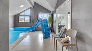 Un espace piscine intérieur moderne, doté d’un toboggan bleu, d’un banc et offrant une vue sur l’extérieur.