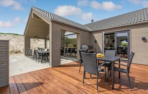 A modern house with a wooden deck, outdoor dining area, and a kitchen visible through glass doors. - Amazing home in Hvide Sande with sauna (Hvide Sande)