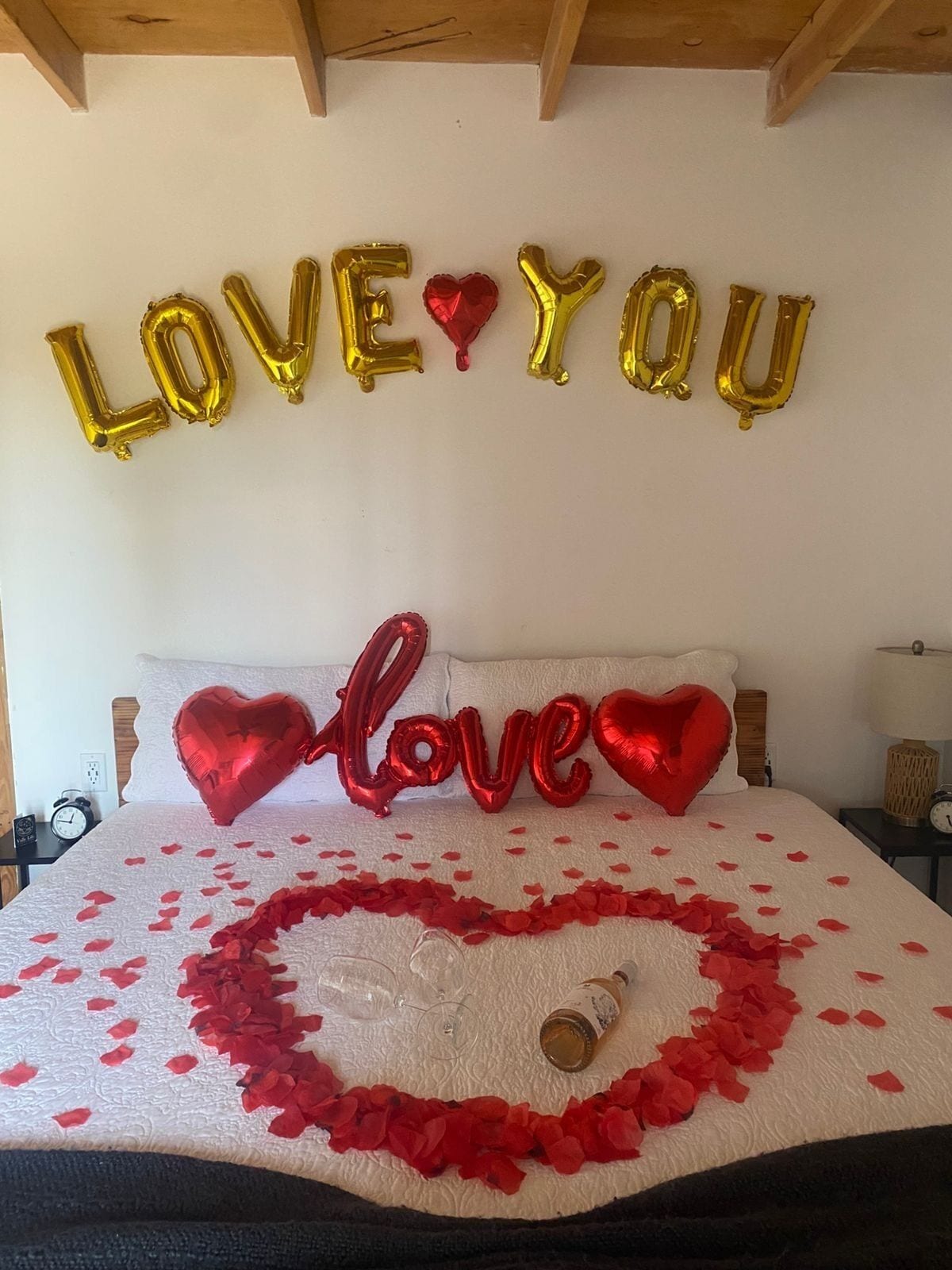 A bed with a heart-shaped arrangement of rose petals and a bottle of wine, surrounded by gold and red balloons spelling 'LOVE YOU'.