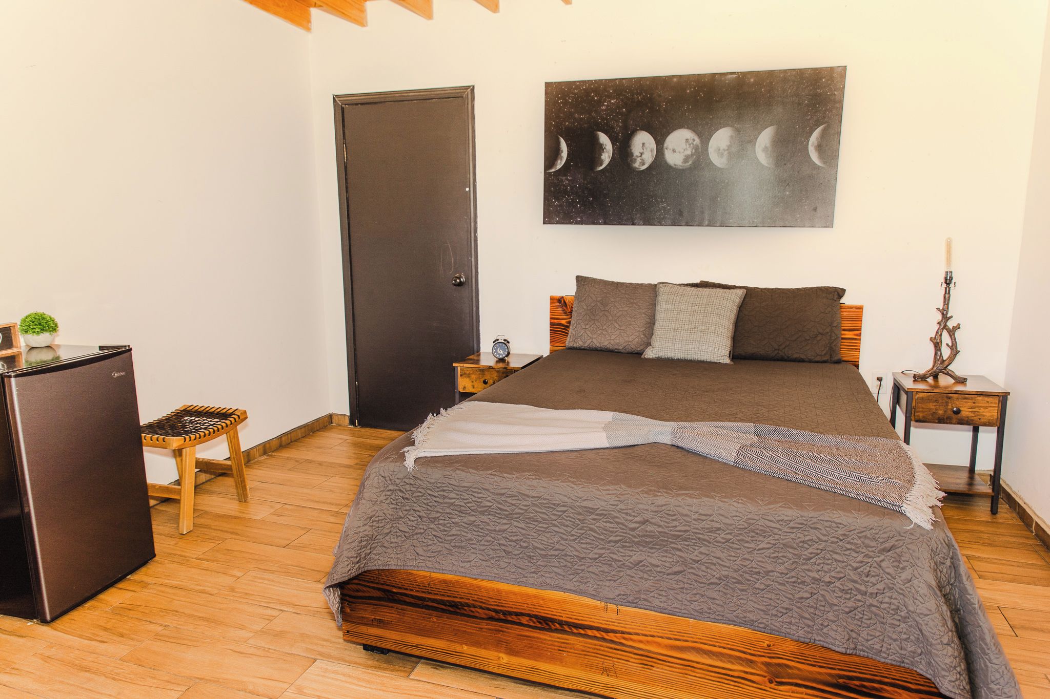 A bedroom with a bed, a nightstand, a lamp, a door, and a wall with a moon phases painting.