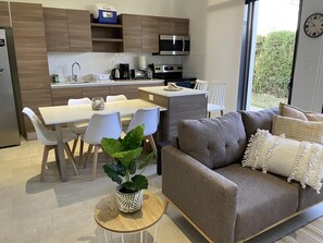 A modern kitchen with a microwave, coffee maker, and toaster. There is a dining area with a table and chairs, and a gray sofa with decorative pillows. A potted plant is on a small table in the center of the room.