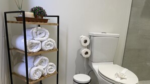 A bathroom with a toilet, a trash bin, and a shelf with rolled towels.