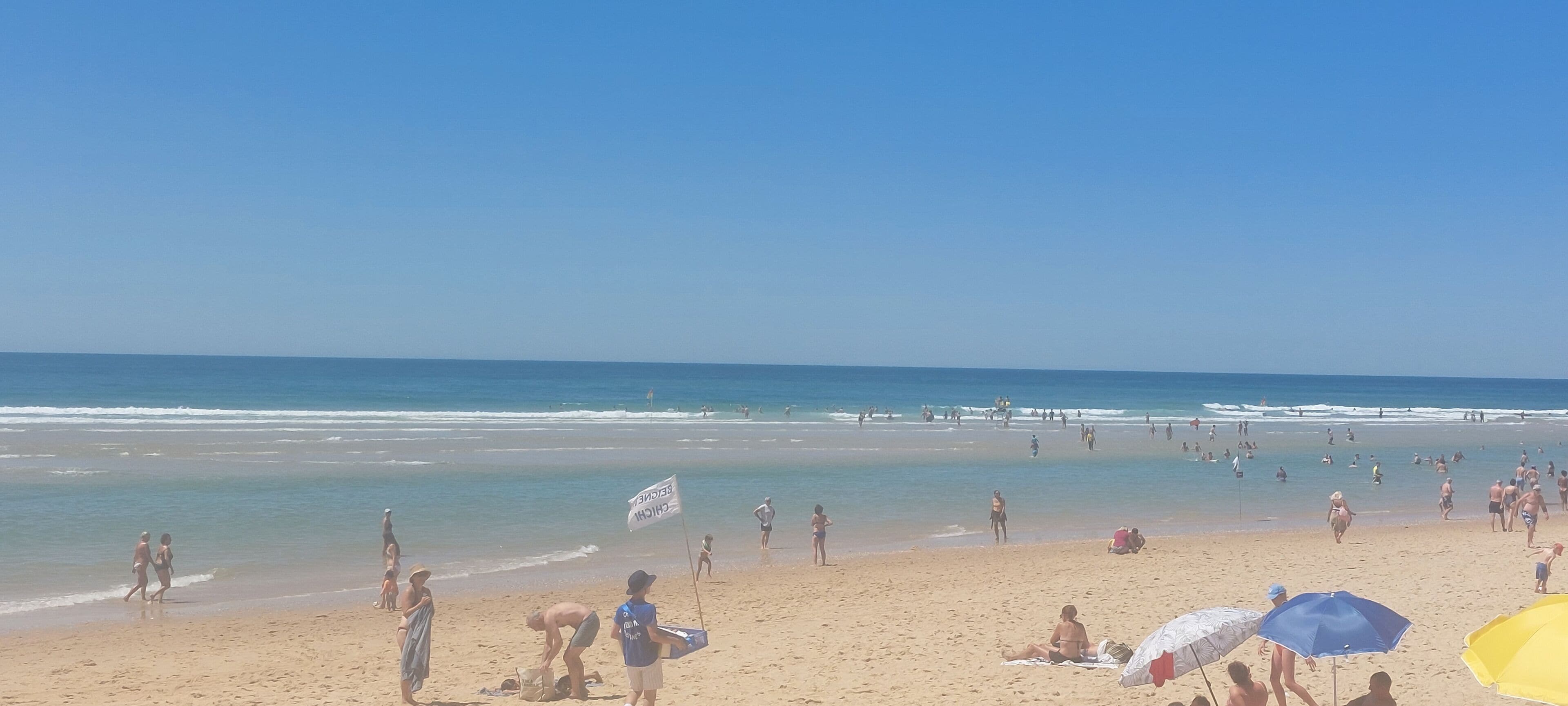 A beach with people walking and sitting on the sand, a lifeguard flag, and umbrellas.