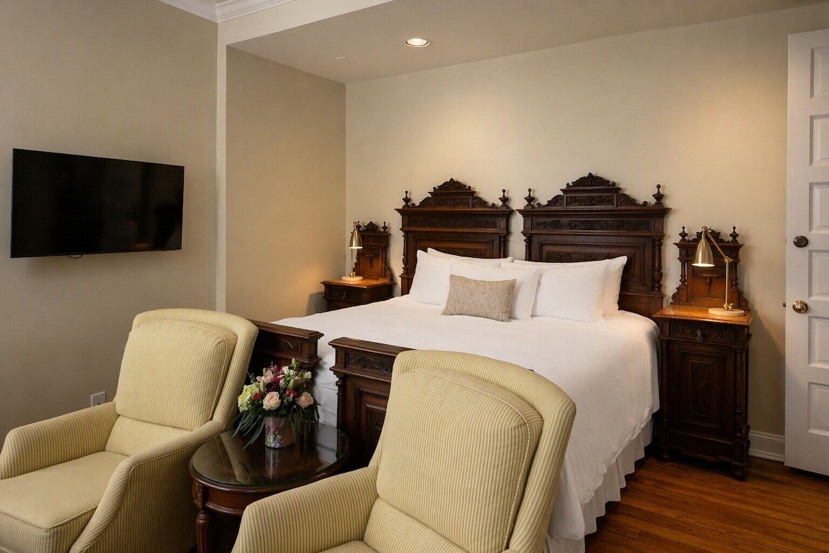 A hotel room with a large bed, two bedside tables with lamps, a television on the wall, and two upholstered chairs.