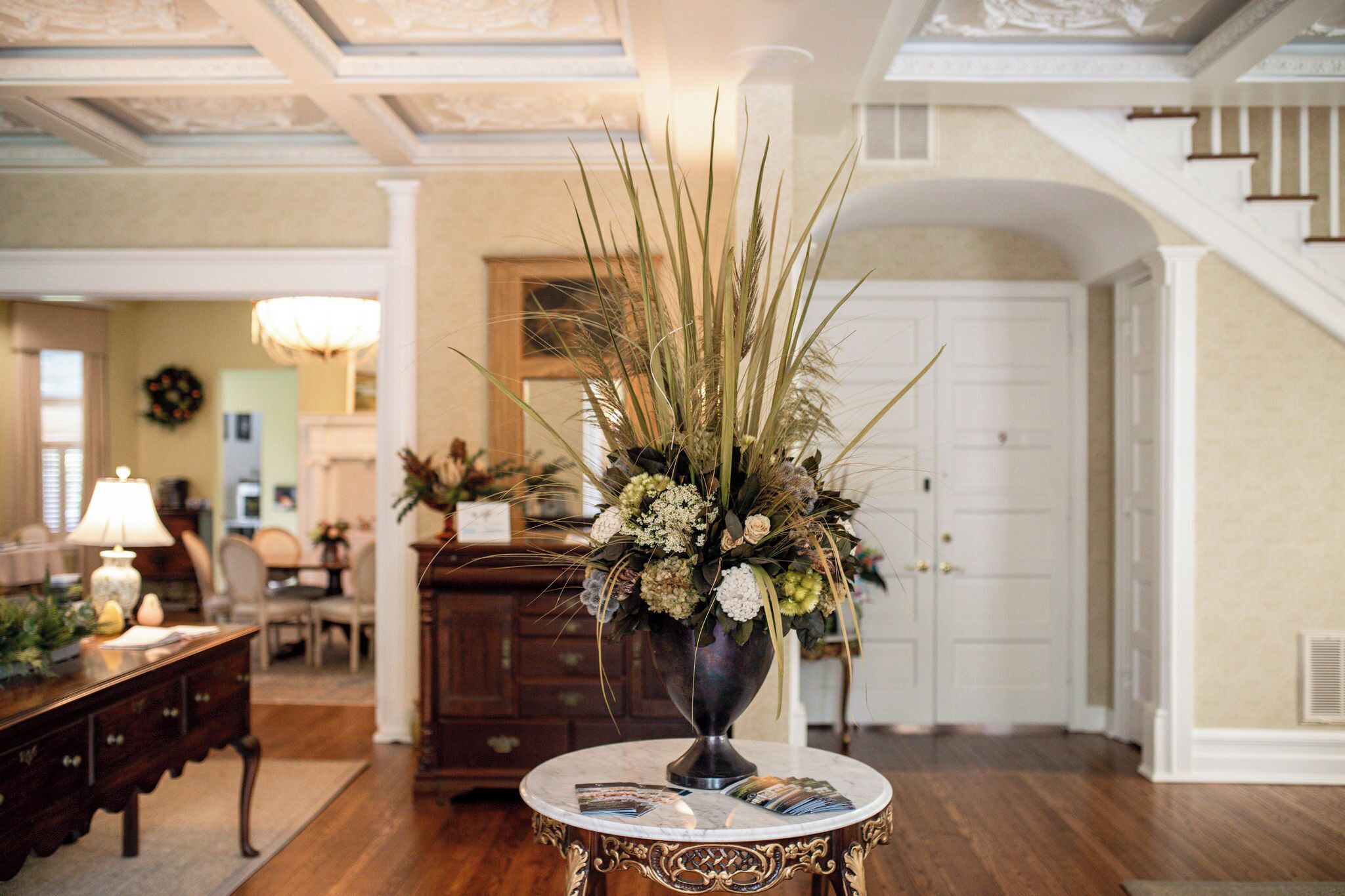 Una habitación con una mesa redonda, un florero con flores, una lámpara, una chimenea y una escalera.