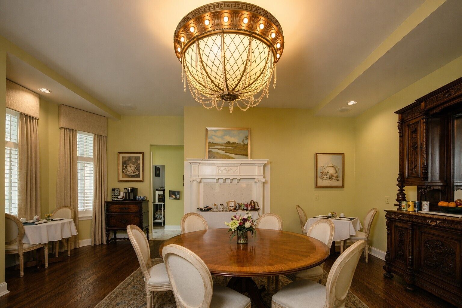 A dining room with a large round table, chairs, a chandelier, a fireplace, and a kitchen area with wooden cabinets.