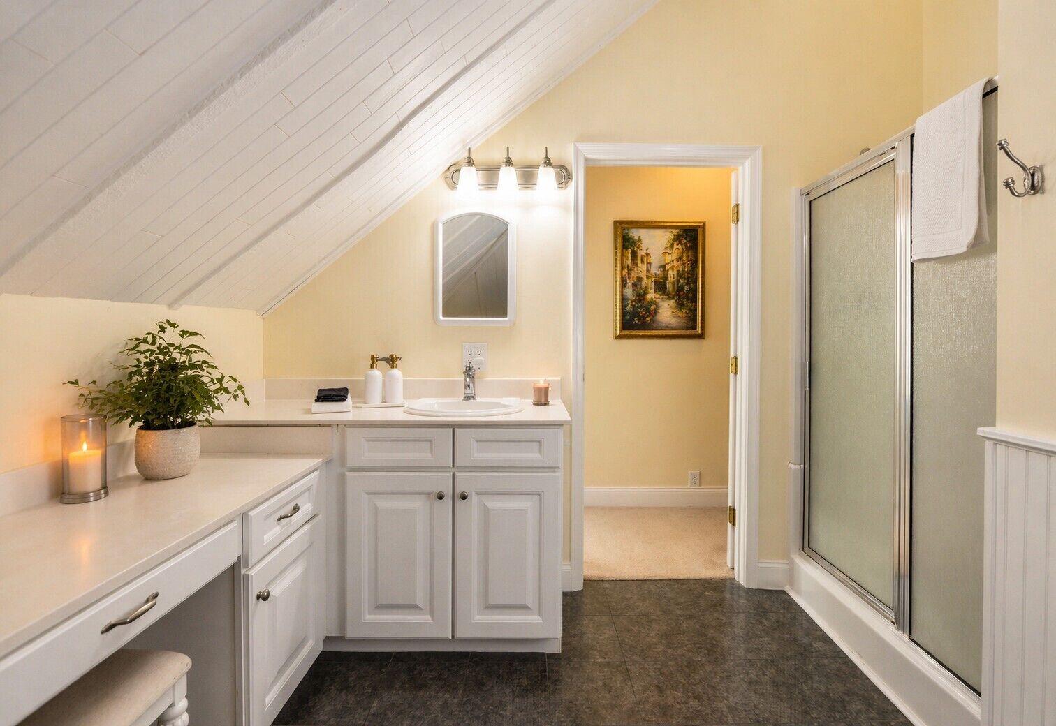 A bathroom with a white vanity, a mirror, a sink, a towel rack, a shower with a glass door, and a painting on the wall.