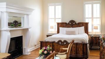 A bedroom with a large bed, a fireplace, a table with chairs, and two windows.