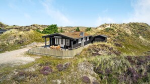 Une maison noire avec une terrasse en bois et un petit abri de jardin, entourée de dunes herbeuses et d’une végétation clairsemée.