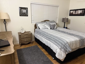 A bedroom with a bed, bedside tables, a desk, a chair, a window with blinds, and wall-mounted pictures.