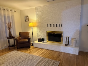 Una sala de estar con chimenea de ladrillo blanco, un sillón de cuero marrón, una lámpara de pie y una mesita con un florero.