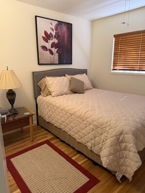 Un dormitorio con cama, mesita de noche, lámpara, un cuadro y ventana con persianas.