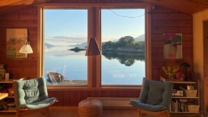 A cabin with two chairs facing a large window overlooking a lake and mountains.