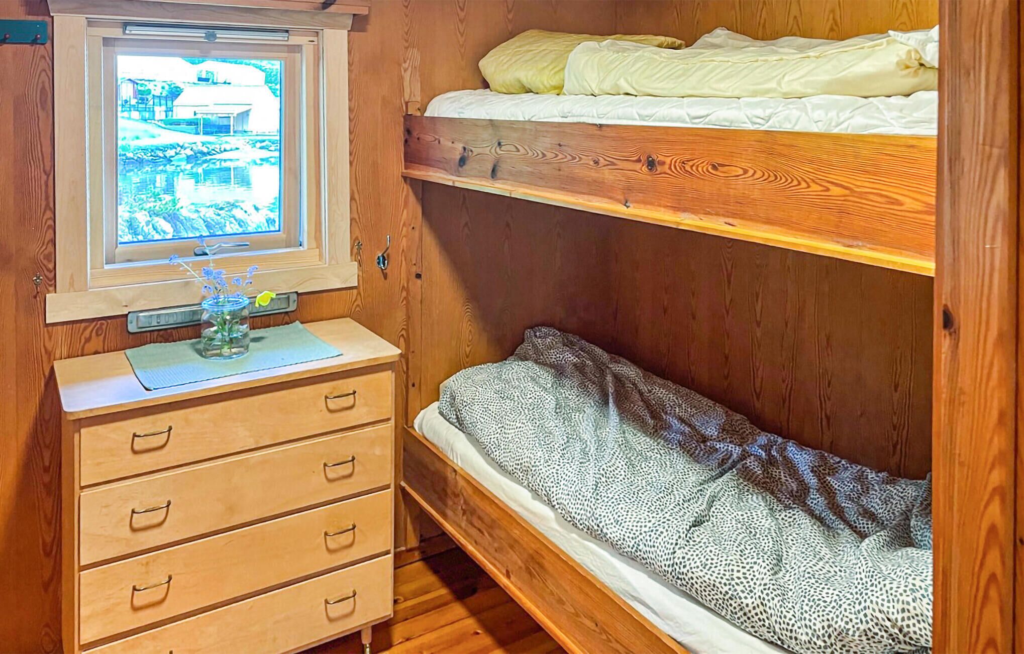 A bunk bed with a window above the lower bunk, a wooden dresser with drawers, and a patterned blanket on the lower bunk.