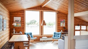 A cozy cabin with wooden walls, a dining table, two blue chairs, and large windows offering a view of the outdoors.
