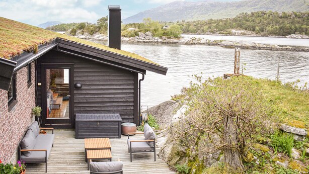 A cabin with a wooden deck, outdoor seating, and a view of a river and mountains.
