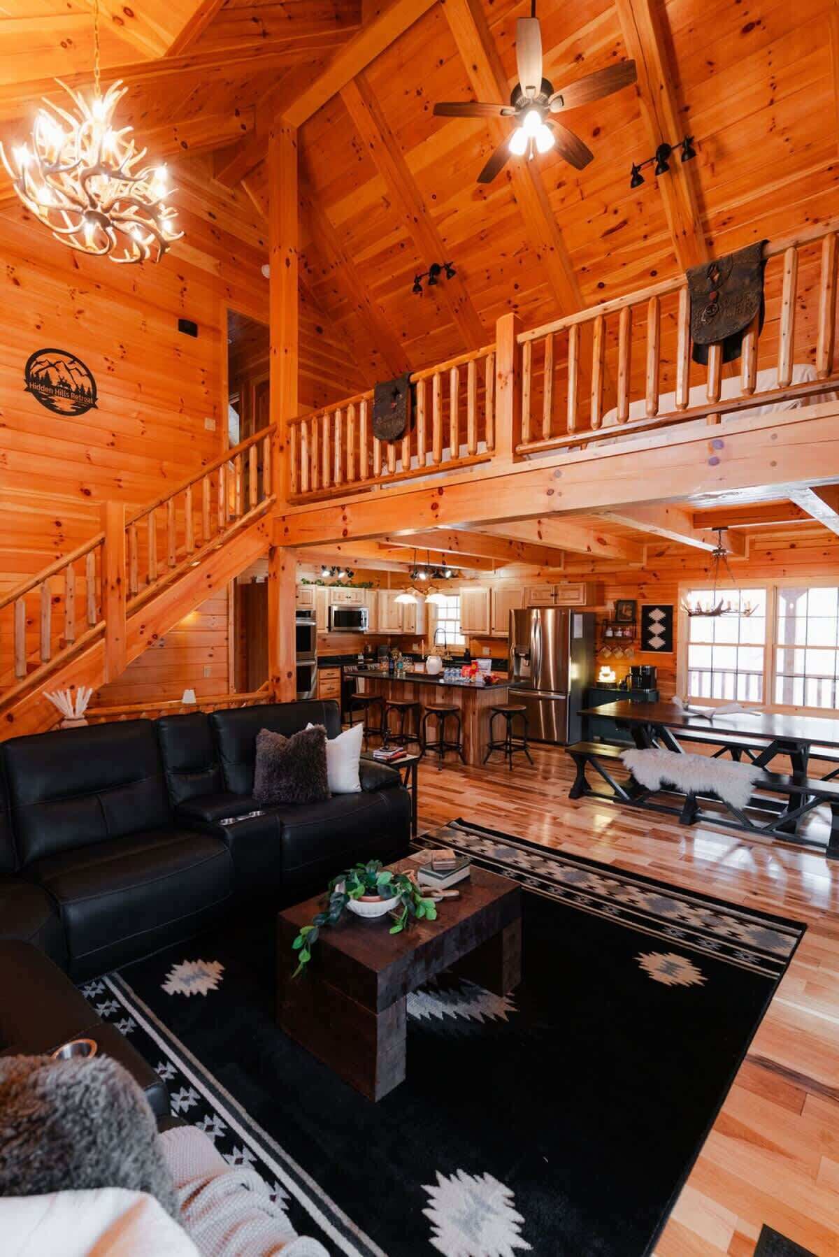 Un intérieur spacieux de chalet en bois, avec un plafond haut, un coin repas, une cuisine et un salon confortable doté d’une cheminée.