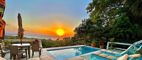 A poolside area with lounge chairs and a table, overlooking a sunset view.