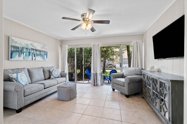 A spacious living room with a grey sofa, a grey armchair, a striped ottoman, and a TV mounted on the wall. - 2 bedroom, walk to the Beach! Sleeps 6, outdoor games and yard space! (Fort Walton Beach)