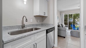 A modern kitchen with a granite countertop, stainless steel sink, and dishwasher. There is a white cabinet above the sink and a blue chair outside the door. - 2 bedroom, walk to the Beach! Sleeps 6, outdoor games and yard space! (Fort Walton Beach)