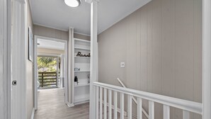 A hallway with a wooden floor, white walls, and a white railing. There is a door leading to a balcony with a view of greenery. - 2 bedroom, walk to the Beach! Sleeps 6, outdoor games and yard space! (Fort Walton Beach)