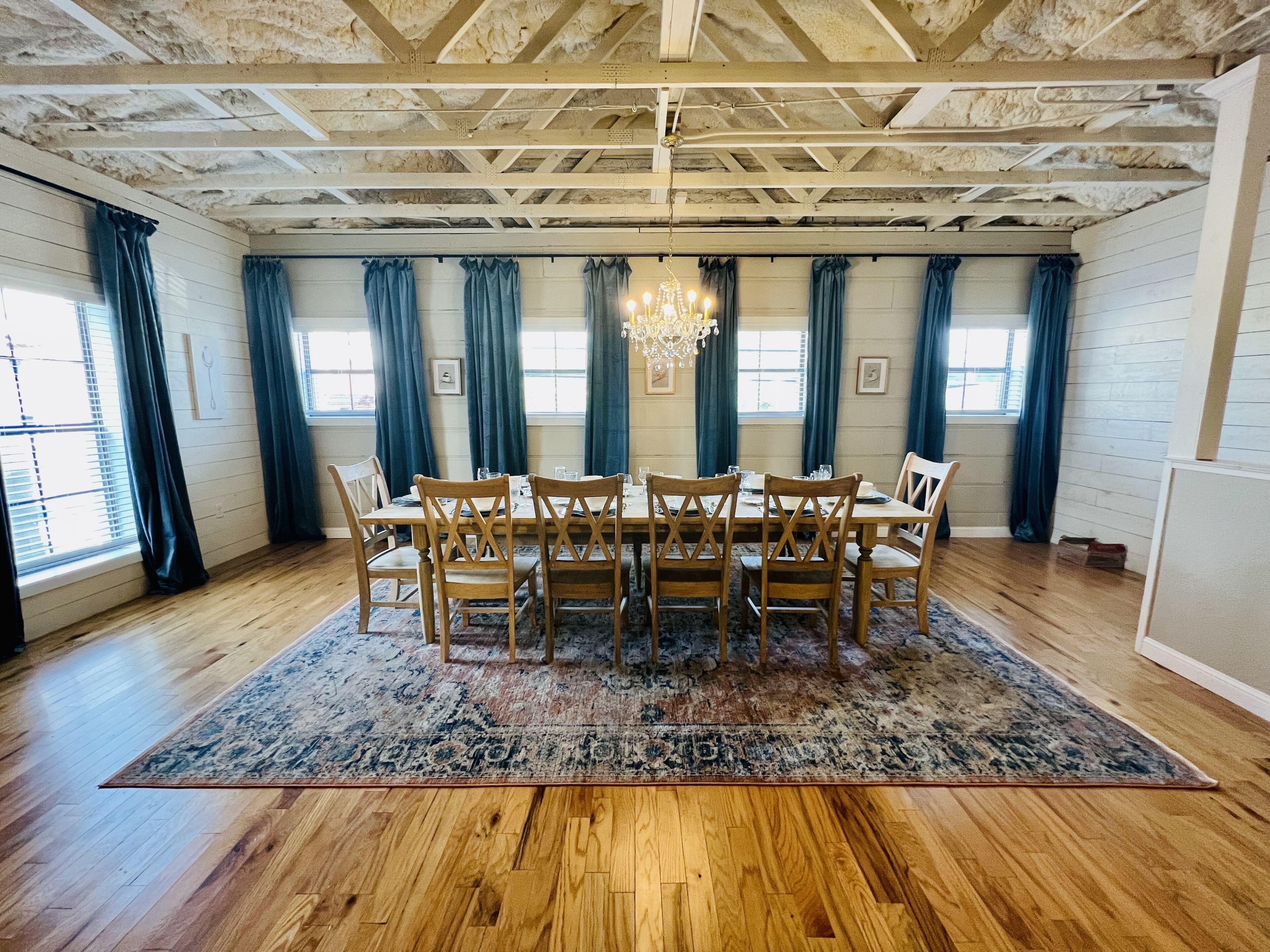 A dining room with a wooden table and chairs, a chandelier, and large windows with curtains.