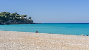 Ein Kiesstrand mit klarem türkisblauem Wasser und einer felsigen Klippe mit Bäumen.
