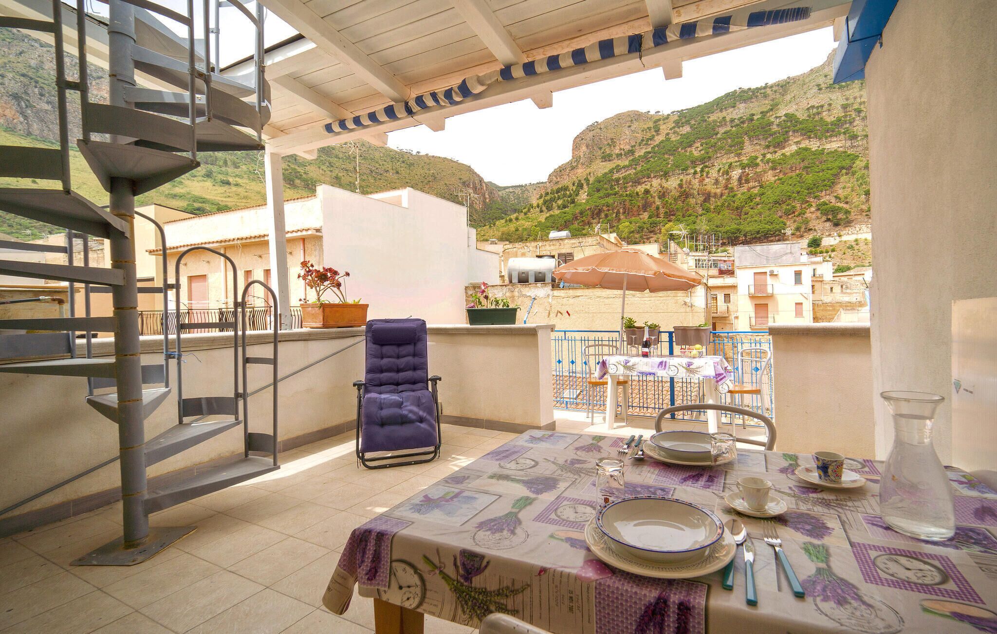 A balcony with a table set for dining, a chair, a potted plant, and a view of a hillside.