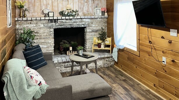 En mysig stuga med en steneldstad, träväggar, en soffa och en tv som är monterad på väggen.
