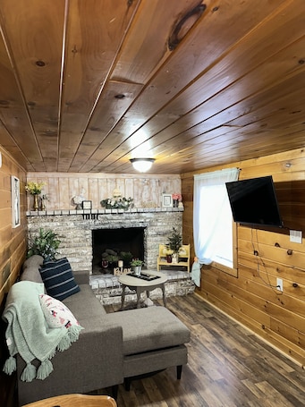 A cozy cabin interior with a stone fireplace, wooden walls, a sofa, and a television mounted on the wall.