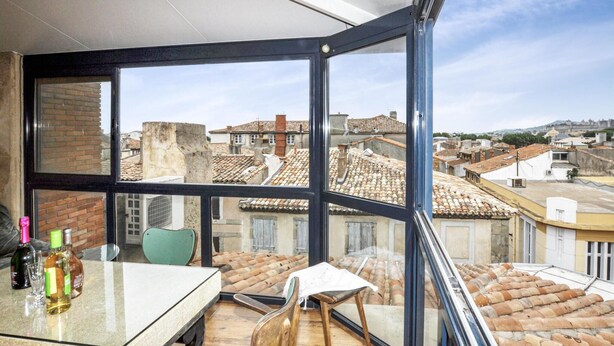 Ein Balkon mit Glastrennwand, einem Tisch mit Flaschen, einem Stuhl und Blick auf Gebäude mit Terrakotta-Dächern.