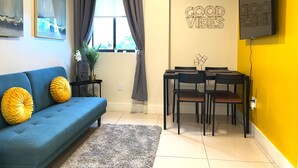 Un salon avec un canapé bleu, une table à manger avec des chaises, un téléviseur fixé sur un mur jaune et des œuvres d’art accrochées aux murs.