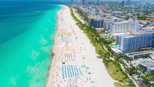 Une vue aérienne d’une ville côtière, avec une longue plage de sable bordée de parasols et un ensemble dense de grands hôtels.