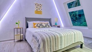 Ein Schlafzimmer mit schrägen Wänden, einem Bett mit gemustertem Überwurf, einem Nachttisch mit Lampe, einem Wanddekor und einem Fenster mit Blick auf Grünflächen.