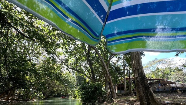 Una balsa de bambú en un río tranquilo, rodeada de exuberante vegetación y un amplio paraguas a rayas que ofrece sombra.