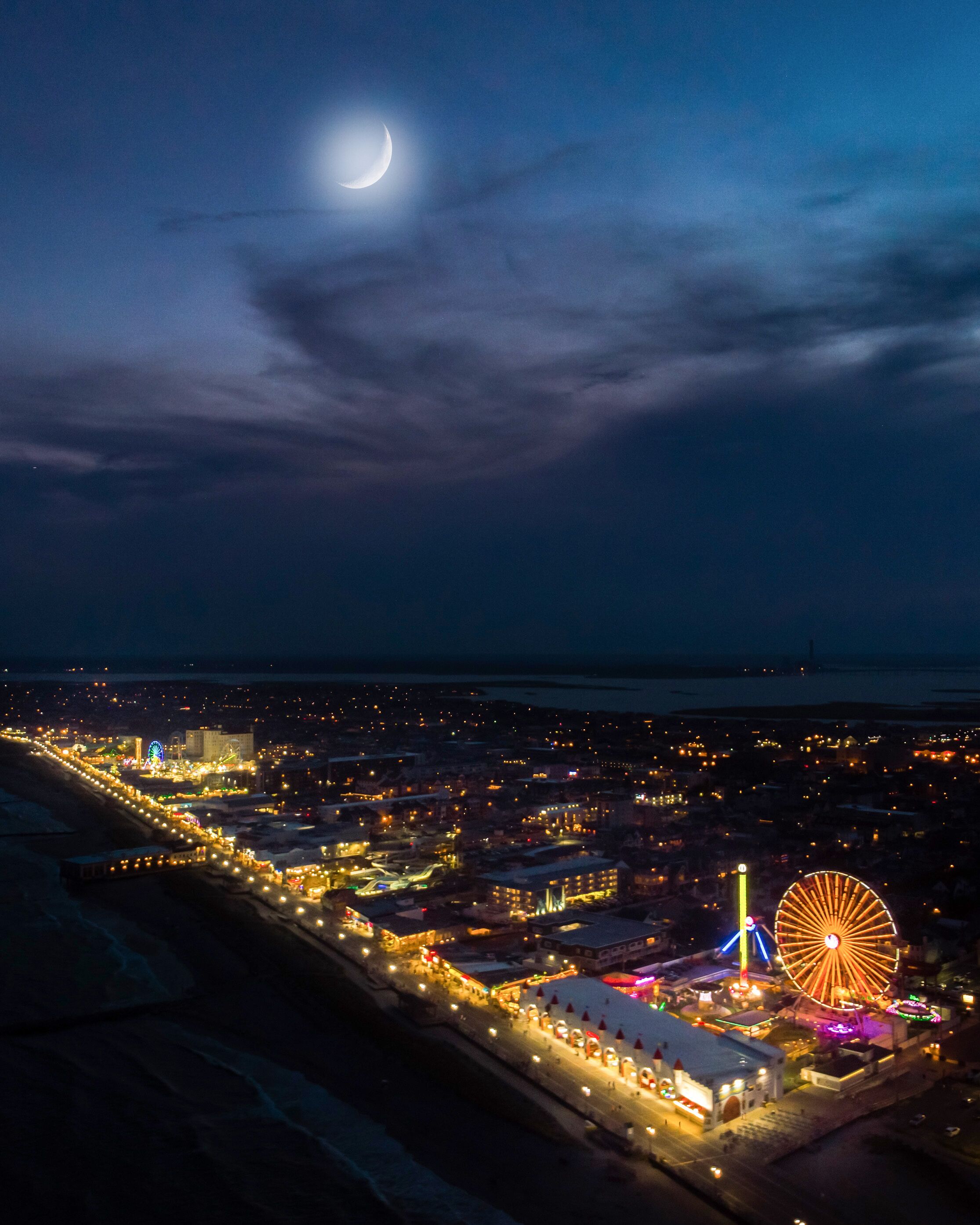 An aerial view of a coastal city at night with a prominent Ferris wheel and a crescent moon in the sky.