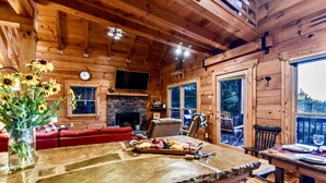 A wooden cabin interior with a stone fireplace, a television mounted on the wall, a dining area with a wooden table and chairs, and a red sofa in the living room. - Spacious Retreat~ Hot Tub~ Stunning Views~ Game Room (Mineral Bluff)