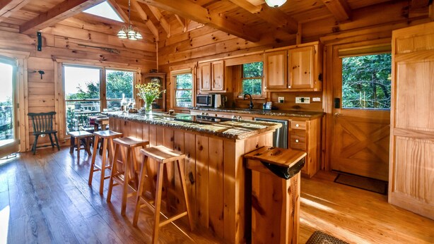 A wooden cabin kitchen with a large island, bar stools, and a view of the outdoors. - Spacious Retreat~ Hot Tub~ Stunning Views~ Game Room (Mineral Bluff)
