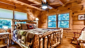 A rustic wooden cabin bedroom with a bed made of logs, a ceiling fan, and a large window with a view of trees. - Spacious Retreat~ Hot Tub~ Stunning Views~ Game Room (Mineral Bluff)