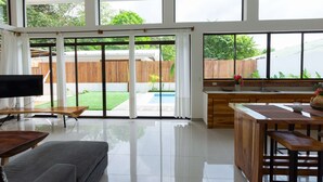 A spacious living area with a large sliding glass door leading to a backyard, a kitchen with a wooden countertop, and a grey sofa. - Villa Koi - Modern and Sophisticated Villa close to the Beach (Santa Teresa beach)