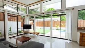 A spacious living room with a large TV, grey sofas, a wooden coffee table, and a view of a backyard with a pool. - Villa Koi - Modern and Sophisticated Villa close to the Beach (Santa Teresa beach)
