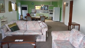 A living room with a sofa, a coffee table, and a dining area with a table and chairs. A kitchen is visible in the background. - Spacious Garden Cottage w/ woodburner (Waitara)