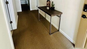 A hallway with a brown carpet, a wooden table with a mirror, and a door leading to another room.