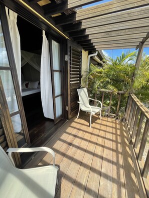 A wooden deck with a white chair, a canopy bed, and a view of palm trees.