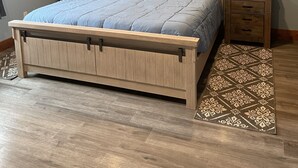 Quarto com cama de madeira, duas mesas de cabeceira, ar-condicionado fixado na parede e um pendente de macramê decorativo.
