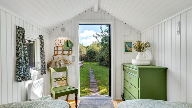 Una habitación con techo de madera, una silla verde, una cama con colcha estampada y un aparador, con vista a un jardín a través de un vano.