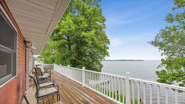 Una terraza de madera con mobiliario exterior, barandilla blanca y vista a un lago.